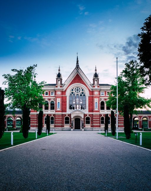 The image shows a large, historic red-brick building with ornate architectural details, including pointed towers, arched windows, and decorative stonework. The building is set against a partly cloudy sky during daylight hours, with the front entrance directly visible and accessible via a paved driveway. Flanking the driveway are symmetrical green lawns with evenly spaced trees providing a balanced landscape. The scene appears calm and well-maintained, suggesting it may be a public or institutional structure. The setting offers a clear view of the building's façade, which is surrounded by the greenery of the trees and open space. This type of building could be associated with a civic, educational, or cultural purpose, and is relevant to house removals and moving services that involve planning and transporting large, historic properties or similar structures, as highlighted by Removal Companies Dulwich in their East Dulwich house removals guide for Lordship Lane, DULWICH.