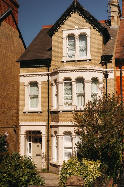 A quiet residential street featuring a narrow, slightly curved asphalt road lined with mature trees and brick boundary walls. In the background, a traditional three-storey red brick house with chimney stacks and sash windows is visible, surrounded by a front garden with shrubs and plants. On the left side, a lamppost stands near the pavement, while on the right side, a black wheelie bin is positioned beside the boundary wall. The scene appears to be late autumn or early winter, with bare tree branches and subdued lighting. This image exemplifies a typical setting for house removals in East Dulwich, with [COMPANY_NAME] often assisting with home relocation, furniture transport, and packing processes to facilitate a smooth move for clients, as highlighted in the East Dulwich house removals guide for Lordship Lane, DULWICH.
