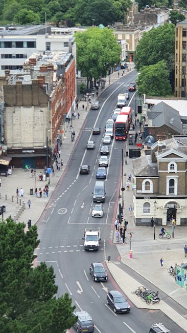 An aerial view of a city street during daytime with multiple vehicles, including cars, vans, and buses, moving along the road. Pedestrians are walking on the sidewalks, which are shaded by green trees lining the street. There are various buildings on both sides, including a brick structure and a small white building with a curved roof, alongside some commercial establishments. The scene captures a typical urban environment that could be part of a house removal or relocation process, with clear road markings and a busy, active setting. This image illustrates the logistics involved in furniture transport and home relocation, highlighting the importance of efficient moving operations managed by a professional removal company such as Removal Companies Dulwich.