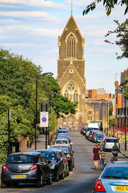 A view of a residential street near Dulwich Picture Gallery in Dulwich, showing a partially visible church with a tall, pointed stone steeple and Gothic-style windows in the background. The street is lined with parked cars on both sides, including small hatchbacks and sedans, with some having visible license plates. Several cardboard boxes, wrapped in brown packing paper and plastic wrap, are stacked on a wheeled trolley inside a property, close to the doorway, ready for home relocation. A person in a red coat and a woman with a bicycle are seen walking along the pavement, possibly transporting belongings. The scene is well-lit with natural daylight, featuring greenery from trees that partly shade the property and street, indicative of a busy but organized local area. The image subtly reflects the logistics of furniture transport and packing involved in professional house removals, with Removal Companies Dulwich performing the loading process as part of their removals service near the historic church and surrounding buildings.