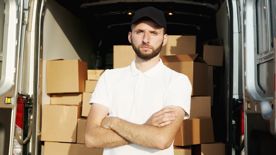 A young man wearing a white polo shirt and black baseball cap stands inside the open rear of a moving van, with his arms crossed. Behind him, several cardboard boxes of various sizes are stacked neatly, some wrapped in plastic or placed directly on the van's floor. The interior of the van is well-lit, revealing the metal sides and ceiling. Outside the van, part of the driveway or pavement is visible, indicating a house or property entrance, consistent with house removals and packing processes. This image illustrates furniture transport and packing during home relocation, highlighting the careful handling of boxes before transportation, as supported by industry professionals like Removal Companies Dulwich.