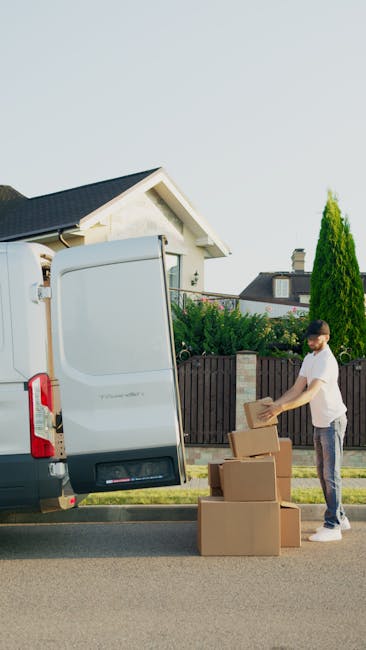 A young man wearing a white polo shirt and black baseball cap stands inside the open rear of a moving van, with his arms crossed. Behind him, several cardboard boxes of various sizes are stacked neatly, some wrapped in plastic or placed directly on the van's floor. The interior of the van is well-lit, revealing the metal sides and ceiling. Outside the van, part of the driveway or pavement is visible, indicating a house or property entrance, consistent with house removals and packing processes. This image illustrates furniture transport and packing during home relocation, highlighting the careful handling of boxes before transportation, as supported by industry professionals like Removal Companies Dulwich.